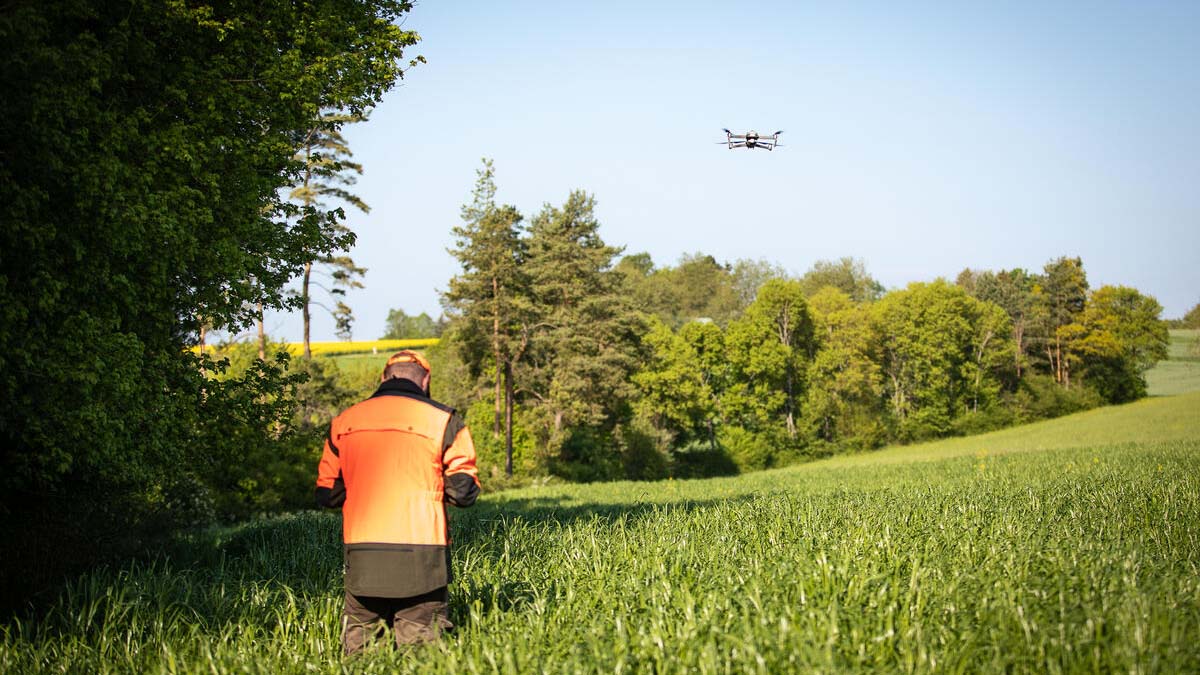 Ein Mann mit orangefarbener Jacke steht in einem Feld. Vor ihm fliegt eine Drohne, welche der Mann steuert.