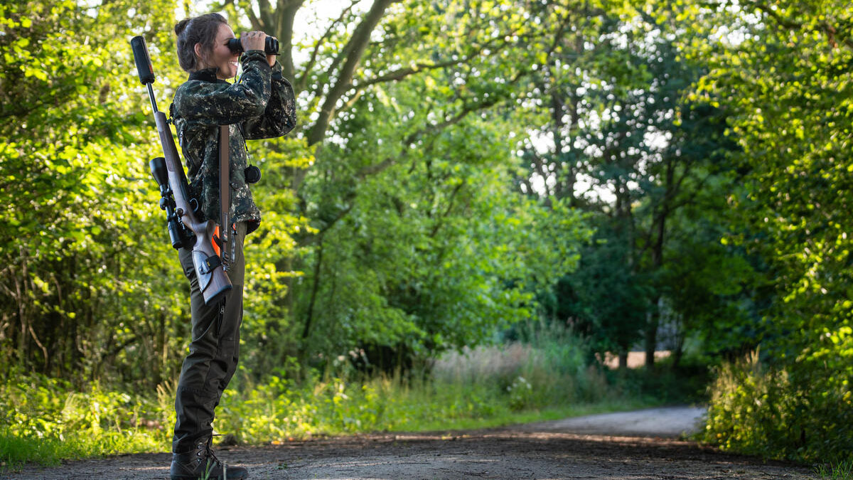 Eine Jägerin steht auf einem Weg im Wald und schaut durch ihr Fernglas. Es ist Sommer.